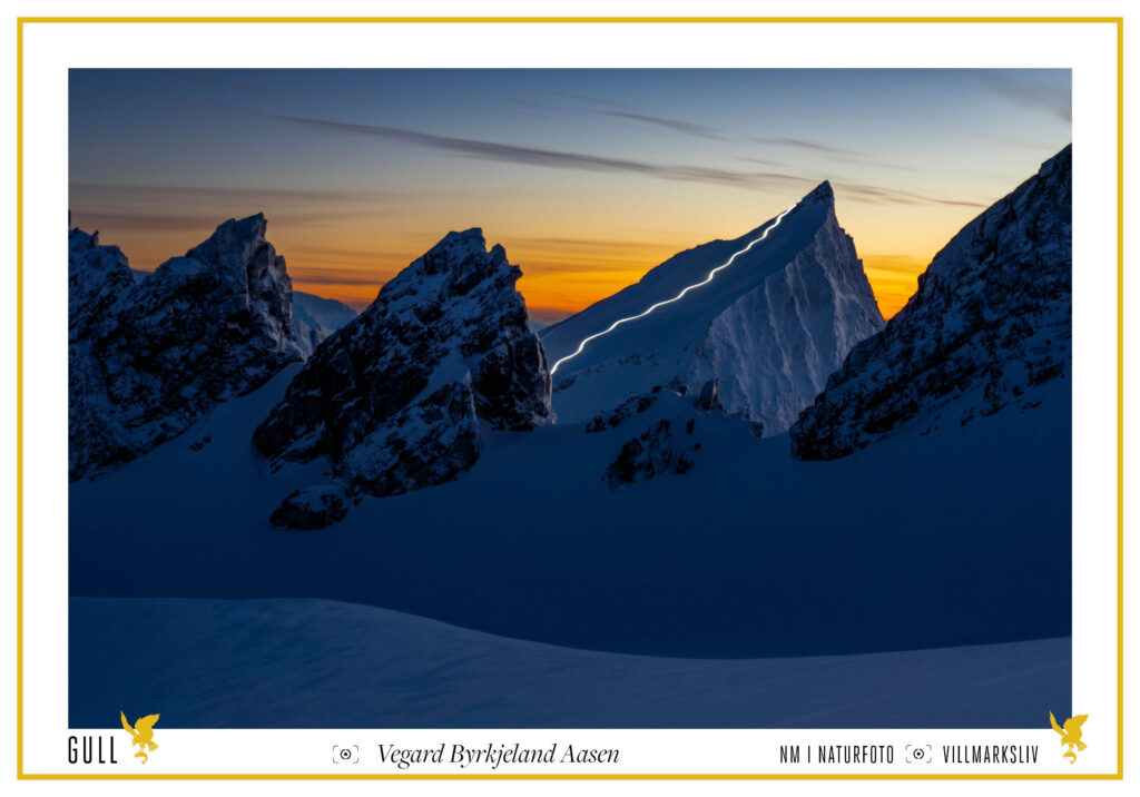 Light Lines 3: Slogen. En skilløper står ned fjellet Slogen med hodelykt. Fotografert på avstand med lang lukkertid, slik at hele fjellsiden lyses opp av en lang lysstripe. Gullbilde i runde 2/2023 i NM i Naturfoto.